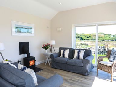 A living room with two gray sofas, a wicker chair, a small wood-burning stove, and large windows with a view at Bwthyn Glanrhyd in Llanfair yn Neubwll