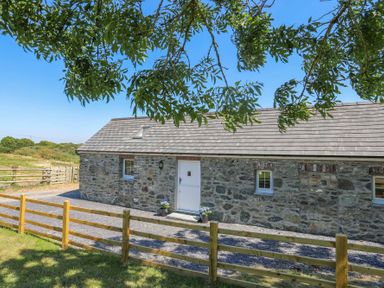 A stone house with white door and windows behind a wooden fence with a tree branch overhead at Bwthyn Glanrhyd in Llanfair yn Neubwll