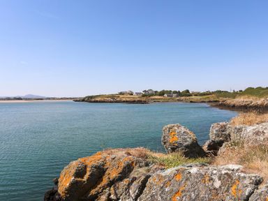 A coastal scene with rocks in the foreground water in the center and houses with vegetation in the background at Bwthyn Glanrhyd in Llanfair yn Neubwll