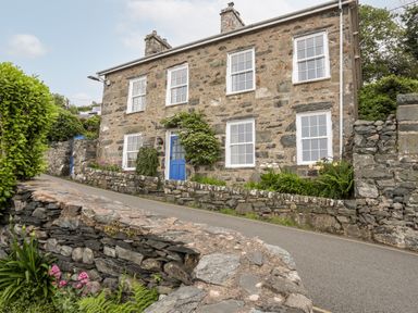 A stone house with a blue door and white framed windows by a paved road and stone walls at Bron Castell in Harlech