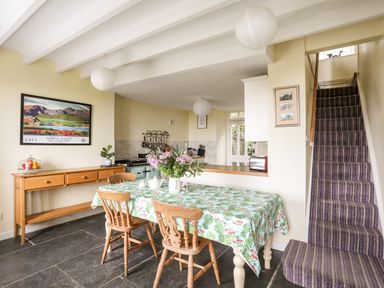 A dining area with a table covered by a patterned tablecloth and wooden chairs near a kitchen and a staircase with striped carpet at Bron Castell in Harlech