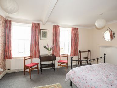 A bedroom with two windows with red curtains two wooden chairs and a small dark wooden table with flowers at Bron Castell in Harlech