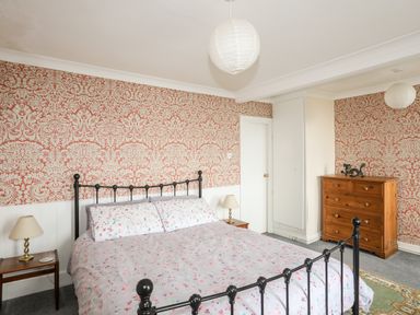 A bedroom with a metal bed frame and floral bedding two bedside tables with lamps a wooden chest of drawers and patterned wallpaper at Bron Castell in Harlech
