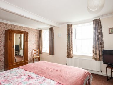 A bedroom with a bed covered in floral and pink bedding a wooden wardrobe with a mirror a wooden chair patterned curtains and a television on a stand at Bron Castell in Harlech