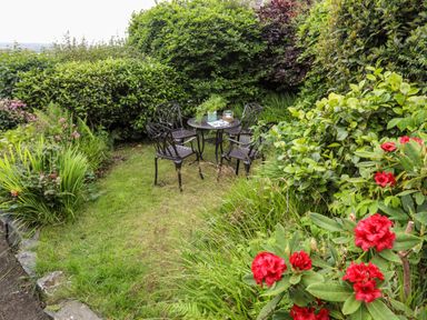 A garden area with a metal table and four chairs surrounded by green bushes and red flowers at Bron Castell in Harlech