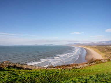 A coastline with waves and a sandy beach beside grassy land and mountains in the background at Bron Castell in Harlech