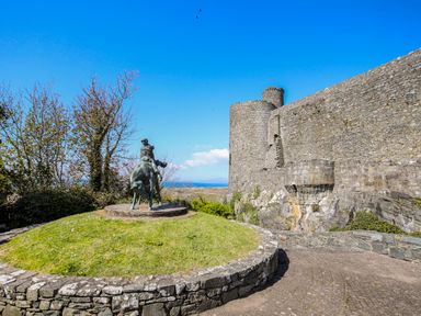 A stone castle wall with a round tower and a statue on a grassy mound in front at Bron Castell in Harlech