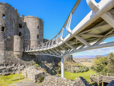 A stone castle with round towers connected by a modern white metal bridge at Bron Castell in Harlech