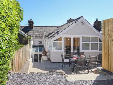 A patio area with a table and six chairs outside a house with white walls and a sloped roof at Brook Cottage in Criccieth
