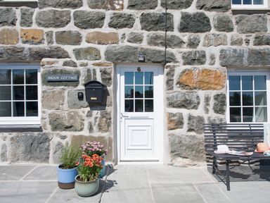 Stone cottage exterior with white door and windows potted plants and a bench at Brook Cottage in Criccieth