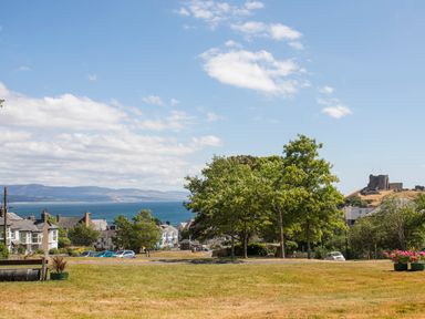 A park with trees and grass overlooking houses by the sea with a castle on a hill in Criccieth