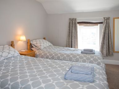 A bedroom with two single beds grey patterned bedding bedside table with lamp window with curtains and mirror on the wall at Brook Cottage in Criccieth
