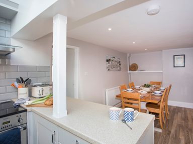 A kitchen counter with two mugs and a spoon looking into a dining room with a wooden dining table set for six at Brook Cottage in Criccieth