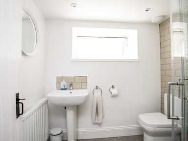 A bathroom with a pedestal sink a toilet a round mirror a towel on a ring and a window at Brook Cottage in Criccieth