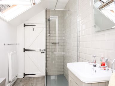 A bathroom with a shower, a sink with faucets and soap, a towel rail, a radiator, and a wooden door at Brook Cottage in Criccieth