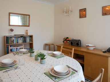 A dining room with a round table set for four with dishes and plants a bookshelf with books a sideboard with a radio flowers and a bowl at Bro Dawel in Beaumaris