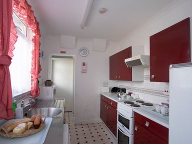 A kitchen with red cabinets a white stove a sink with a basket of bread and eggs and pink curtains at Bro Dawel in Beaumaris