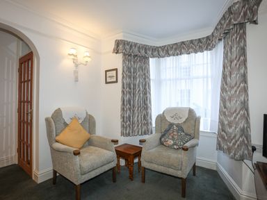 A sitting area with two armchairs and a wooden side table between them in a room with patterned curtains and a glass-paned wooden door at Bro Dawel in Beaumaris
