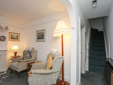 A sitting area with two armchairs, a floor lamp, a table lamp, framed pictures on the wall, and a staircase in a hallway at Bro Dawel in Beaumaris