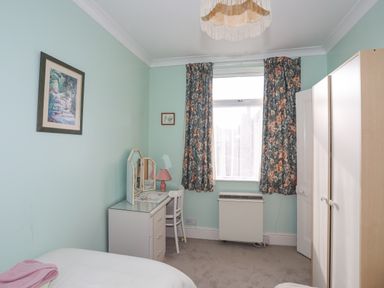 A bedroom with a bed a dressing table with mirror a chair a lamp floral curtains and a wardrobe at Bro Dawel in Beaumaris