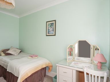 A bedroom with a single bed with pillows and folded towels and a dressing table with a tri-fold mirror and a pink lamp at Bro Dawel in Beaumaris