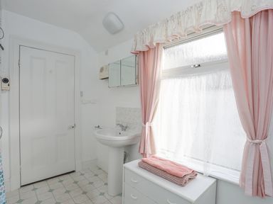 A bathroom with a pedestal sink a window with pink curtains and a white cabinet with folded towels at Bro Dawel in Beaumaris