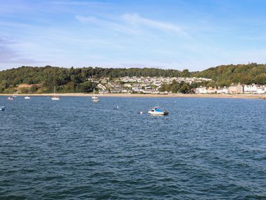 A coastal area with boats on water and houses along a shoreline with trees behind at Bro Dawel in Beaumaris