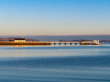 A pier extending over calm water with a small building and hills in the background at Bro Dawel in Beaumaris
