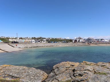 A coastal scene with rocks in foreground a curved pier and buildings along the shoreline at Bryn Dale in Trearddur Bay