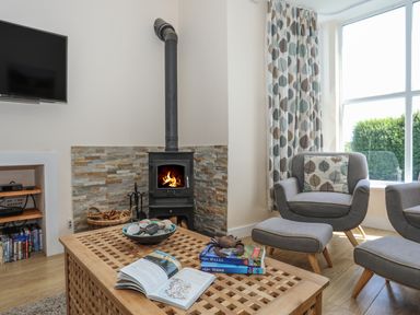 A living room with a wood stove fireplace a wooden coffee table with books and shells grey armchairs with leaf patterned cushions and curtains at Bryn Dale in Trearddur Bay