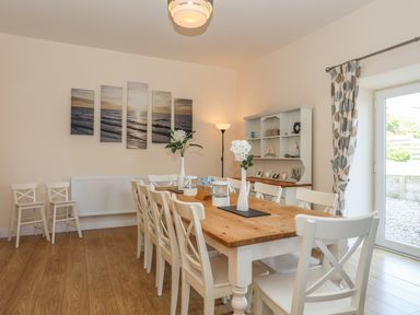 A dining room with a wooden table and white chairs next to a window with patterned curtains at Bryn Dale in Trearddur Bay