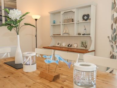 A dining room with a wooden table white chairs a white vase with flowers and a white cabinet with decorative items at Bryn Dale in Trearddur Bay