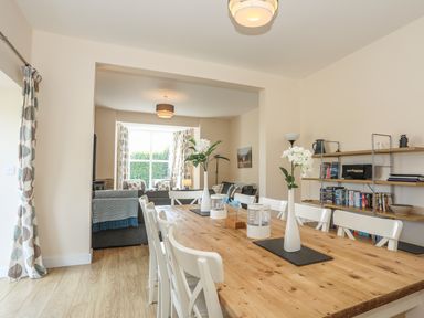 A dining area with a wooden table and white chairs next to a living room with sofas and a large window at Bryn Dale in Trearddur Bay