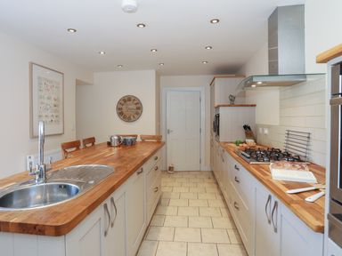 A kitchen with wooden countertops, a sink, a stove, a clock on the wall, chairs at an island counter, and kitchen cabinets at Bryn Dale in Trearddur Bay