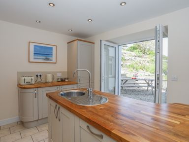A kitchen with wooden countertops a sink and an open door leading to an outdoor picnic table with flowers at Bryn Dale in Trearddur Bay