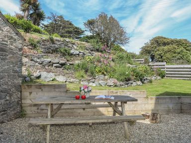 A wooden picnic table with apples a book and flowers in a garden with stone walls and plants at Bryn Dale in Trearddur Bay