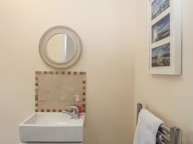 A small bathroom with a square sink a round mirror above it a soap dispenser a towel on a metal towel rack and framed pictures on the wall at Bryn Dale in Trearddur Bay