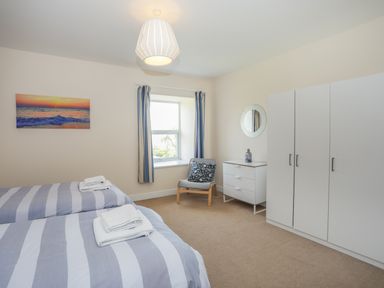 A bedroom with two striped beds a chair a window with curtains a white wardrobe and a chest of drawers at Bryn Dale in Trearddur Bay