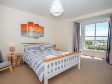 A bedroom with a bed striped bedding two bedside tables with lamps a chair and large window at Bryn Dale Trearddur Bay