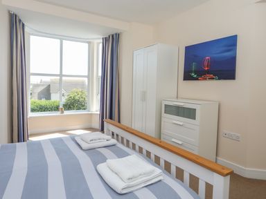 A bedroom with a bed, striped bedspread, folded towels, white wardrobe, chest of drawers, and large window with curtains at Bryn Dale in Trearddur Bay