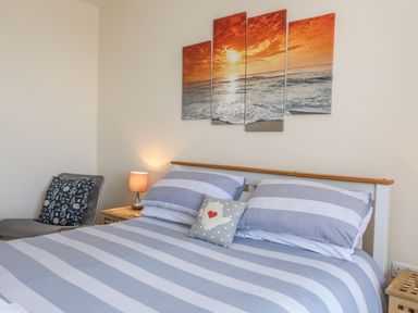 A bedroom with a bed with striped bedding a bedside table with a lamp a chair with a cushion and a four panel sunset ocean wall art at Bryn Dale in Trearddur Bay