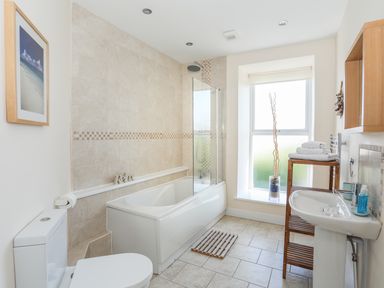 A bathroom with a bathtub shower combination a toilet a pedestal sink wooden shelving with towels and a window at Bryn Dale in Trearddur Bay