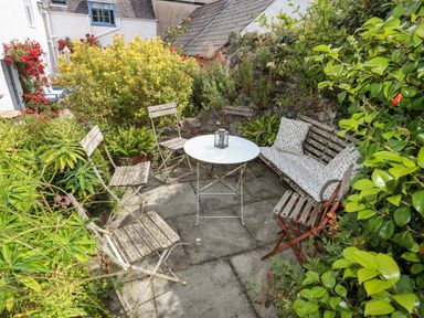 An outdoor seating area with a white round table surrounded by wooden chairs and a bench with cushions in a garden at Bryn Tegid in Beaumaris