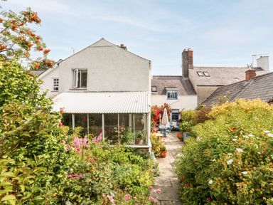 Garden with a paved pathway leading to a white house with a conservatory and outdoor seating area at Bryn Tegid in Beaumaris