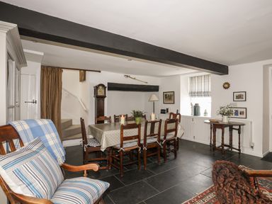A dining room with a wooden table and chairs a grandfather clock a wooden armchair with cushions and a window with a striped blind at Bryn Tegid in Beaumaris