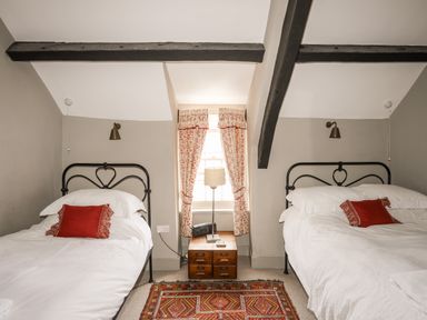 A bedroom with two single beds with red pillows a wooden nightstand a lamp a radio window with floral curtains and exposed ceiling beams at Bryn Tegid in Beaumaris