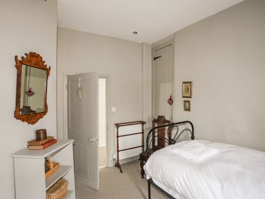 A bedroom with a bed with white linens a wooden chair a small open shelf and a mirror on the wall at Bryn Tegid in Beaumaris
