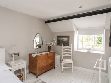A bedroom corner with a wooden chest, white chair, bedside table with lamp and window at Bryn Tegid in Beaumaris