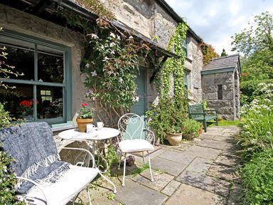 A stone cottage exterior with a small patio table and chairs plants and a green bench at Buck Cottage in Red Wharf Bay