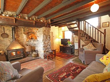 A living room with a stone fireplace a wood burning stove armchairs a sofa rugs and wooden stairs at Buck Cottage in Red Wharf Bay